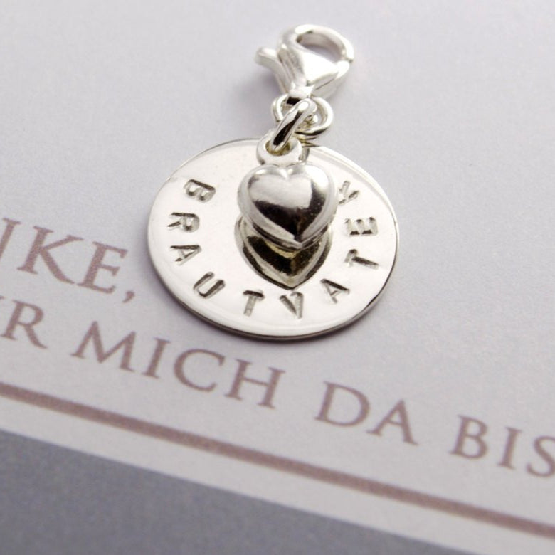 Detailansicht eines Hochzeits-Anhängers als Geschenk für den Brautvater. Wunschgravur auf einem 15 mm Silberplättchen, hier 'BRAUTVATER' über dem mittig ein 925 Silber Herz-Anhänger befestigt ist. Der Charm ist mit einem Karabiner versehen und wird in einer kleinen grau-blauen Geschenkbox verpackt.
