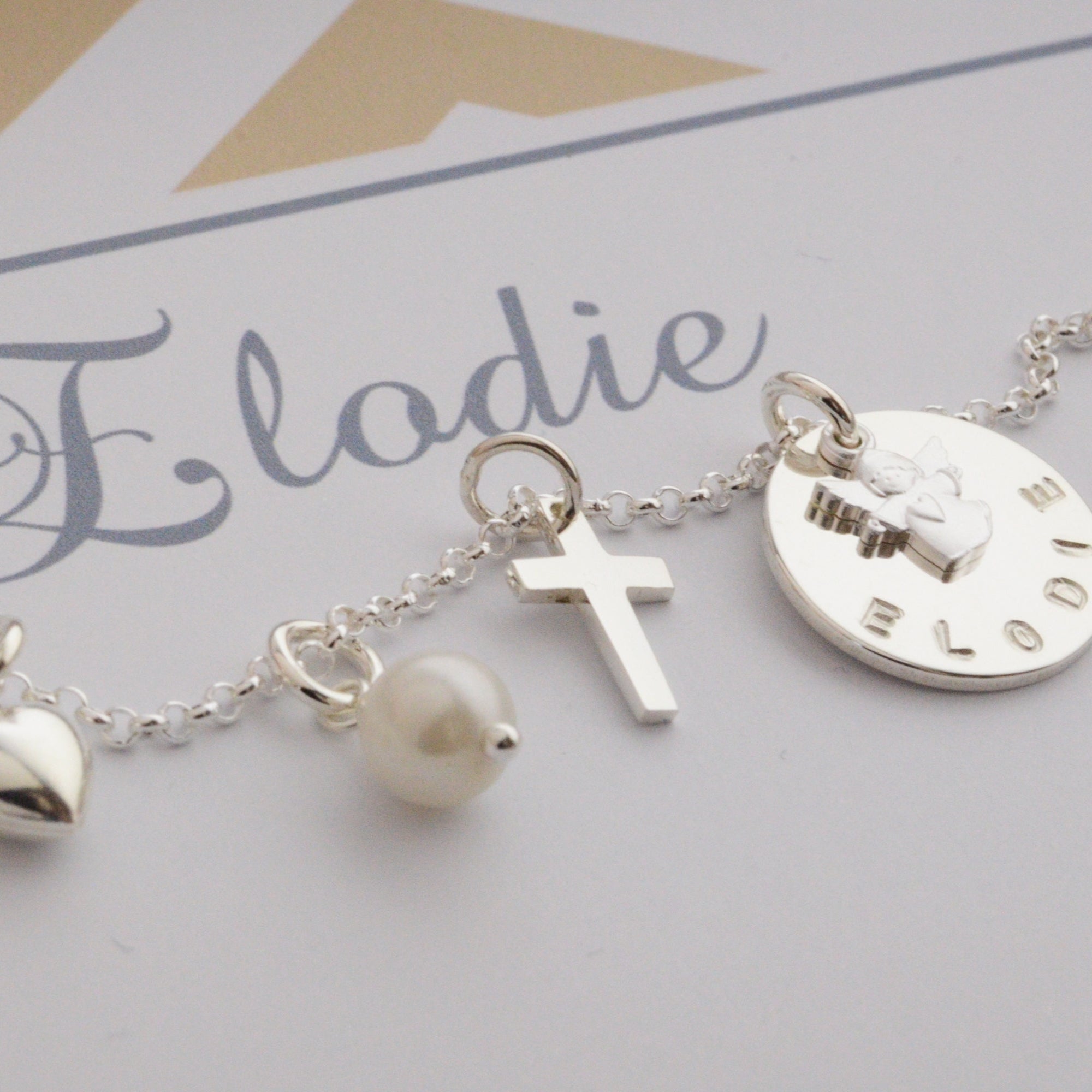 Detailansicht eine Silberkette als Geschenk zur Kommunion mit Herz, Kreuz, Perle Namensanhänger und Schutzengel in einer personalisierten Geschenkbox in beige und weiß.