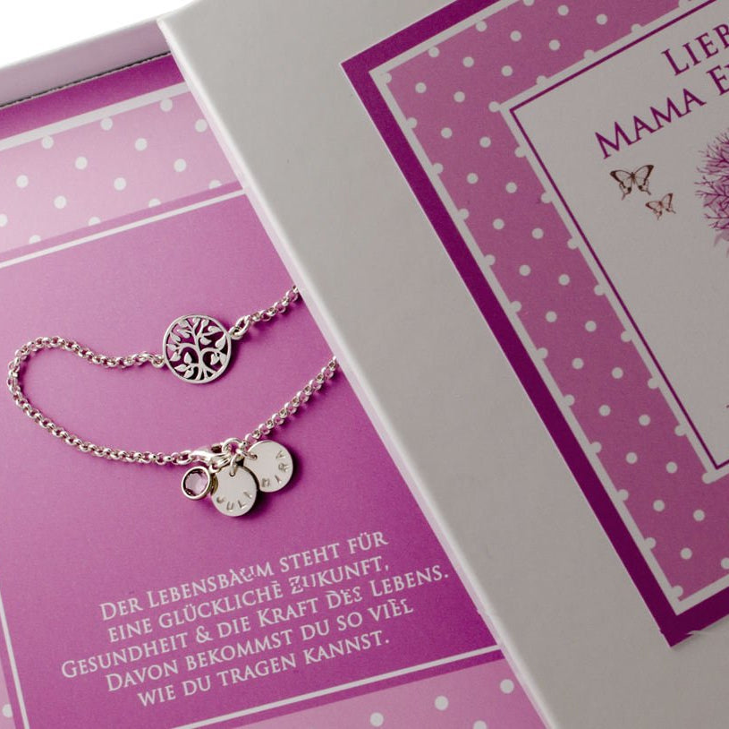 Detailansicht einer Lebensbaum-Armbandes mit zwei Gravur-Anhängern mit Wunschgravur, hier 'JULI' und 'MIRA", präsentiert in einer personalisierten, mit Pünktchen-Muster versehenen Geschenkbox.
