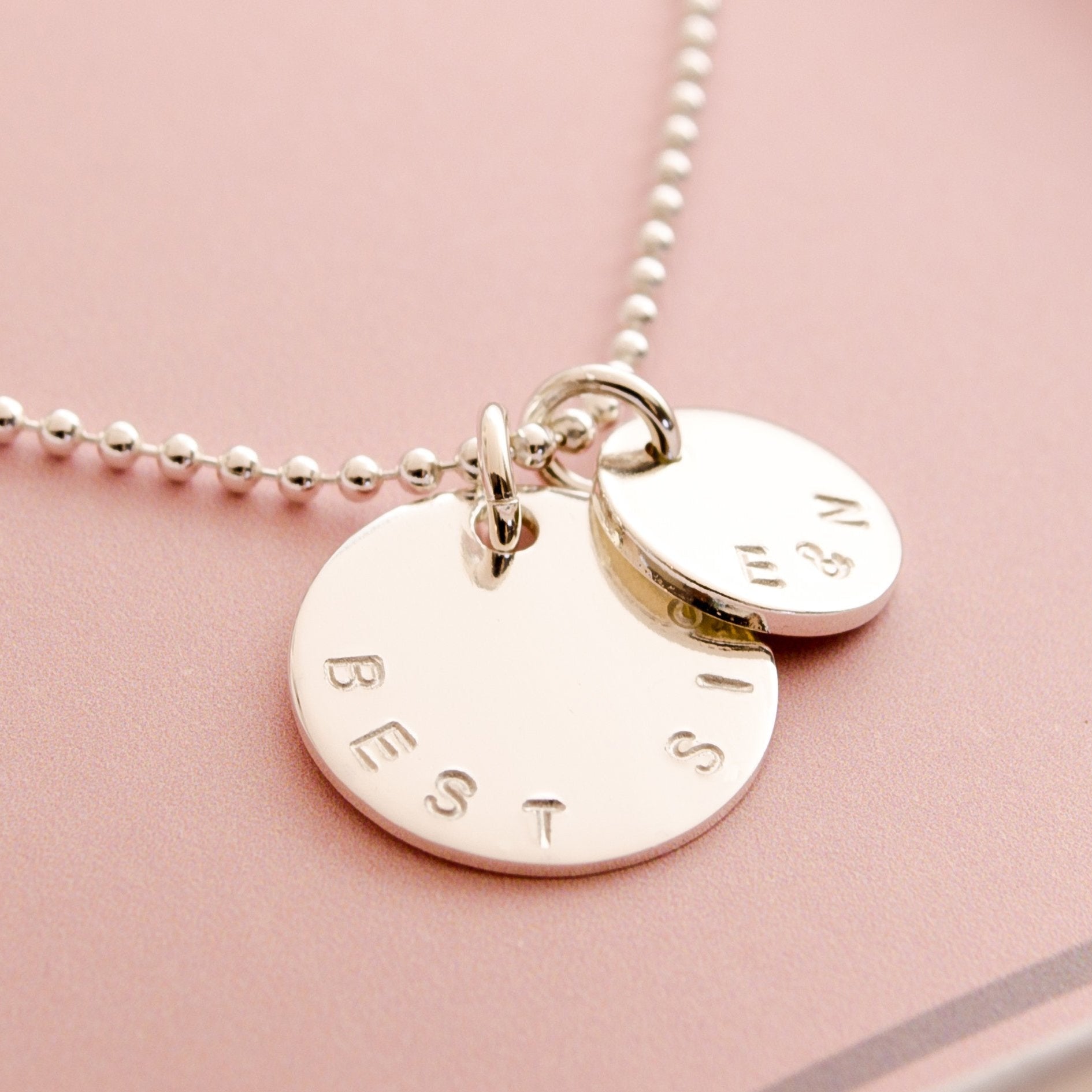 925 Silber Kugelkette mit großem, on Hand gestempeltem Anhänger 'BEST SIS' und kleinem Initial-Taler, hier mit 'E&N' auf einer rosa Geschenkbox abgebildet.