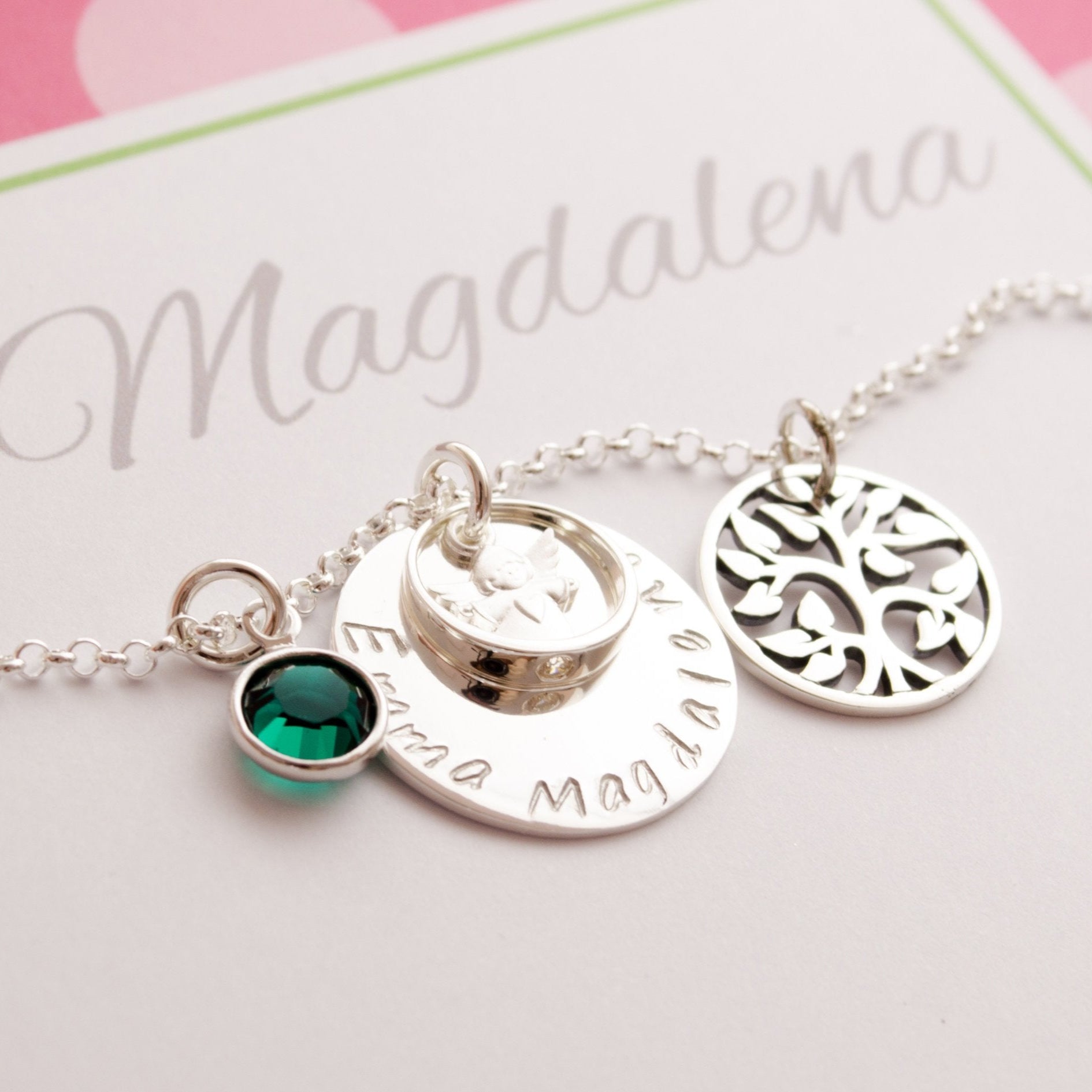 Detailansicht einer taufkette aus 925 Sterlingsilber mit großem Namensplättchen, Engel im Taufring, Lebensbaum und Geburtsstein. Die Kette liegt in einer bunt gepunkteten, personalisierten Geschenkbox.