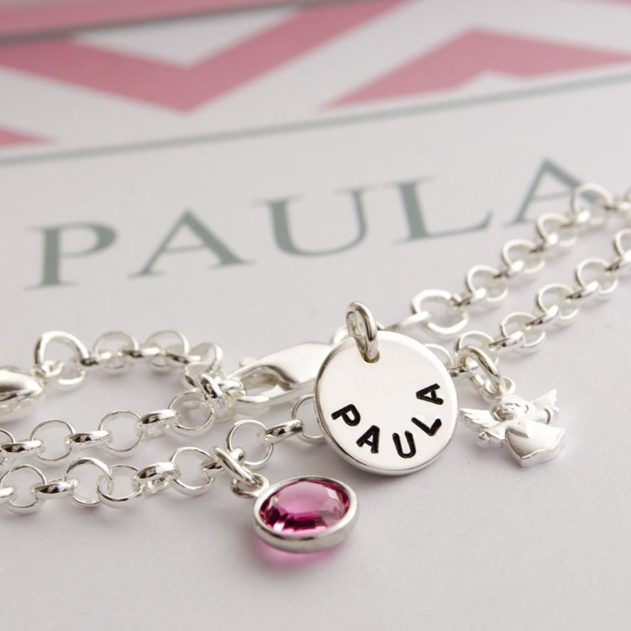 Detailansicht eines Taufarmbandes mit handgestempeltem Namensplättchen 'PAULA' aus 925 Sterling Silber, einem süßen kleinen Schutzengel und Geburtsstein, hier in Oktober dunkel rosé. ads Armband liegt in einer personalisierten Geschekbox mit rosa farbenem Zickzack-Muster, handgefertigt von Bloomgart.