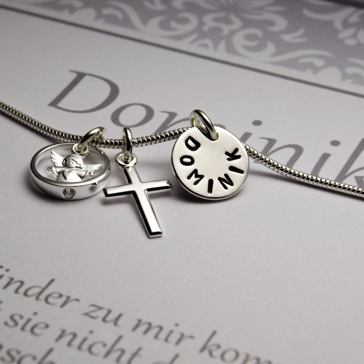 Makroansicht der Taufkette 'Dominik' mit Schutzengel im Taufring, Kreuz und handgestempeltem Namenstaler in einer personalisierten Geschenkbox mit grau-weißem Ornamentmuster präsentiert.