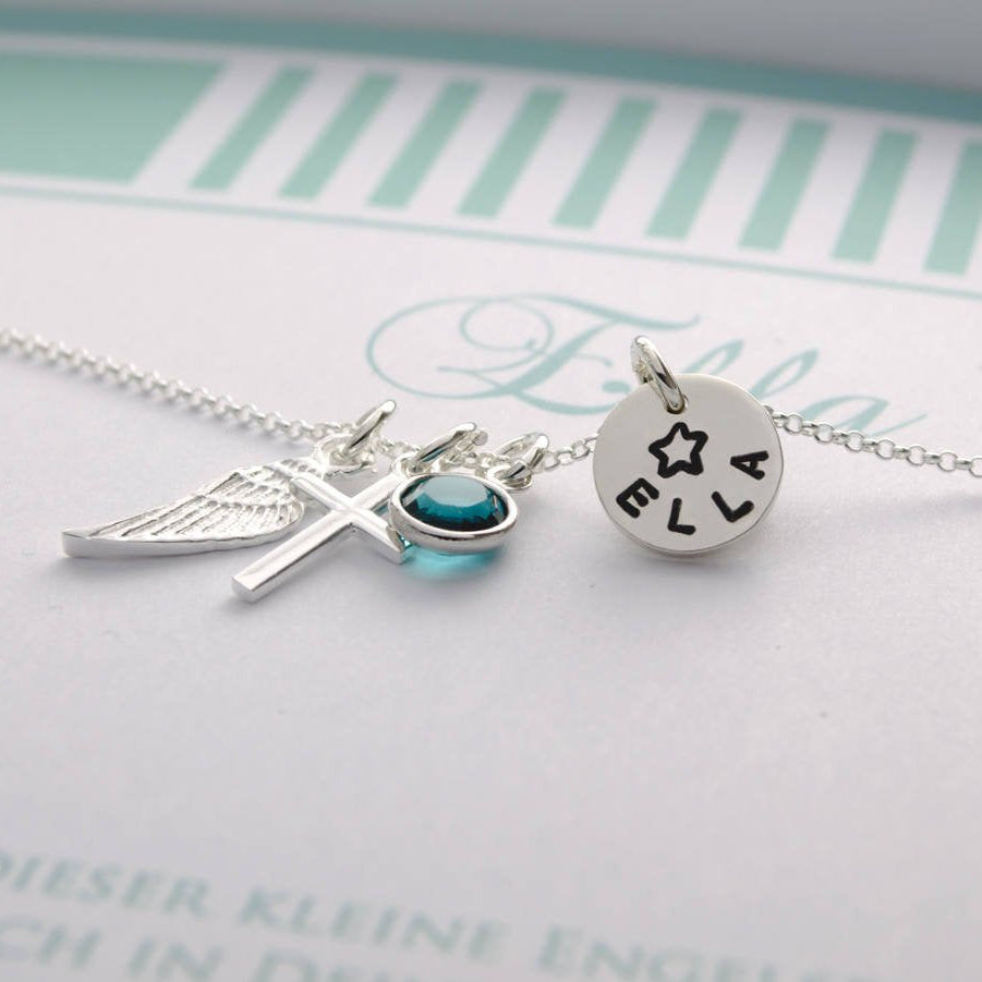 Nahaufnahme einer personalisierten Schutzengel-Taufkette aus 925 Silber. Mit Engelsflügel, Kreuz, blauem Geburtsstein und einem Plättchen, das mit dem Namen 'Ella' und einem Stern graviert ist. Von Bloomgart.