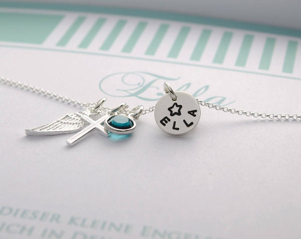 Nahaufnahme einer personalisierten Schutzengel-Taufkette aus 925 Silber. Mit Engelsflügel, Kreuz, blauem Geburtsstein und einem Plättchen, das mit dem Namen 'Ella' und einem Stern graviert ist. Von Bloomgart.