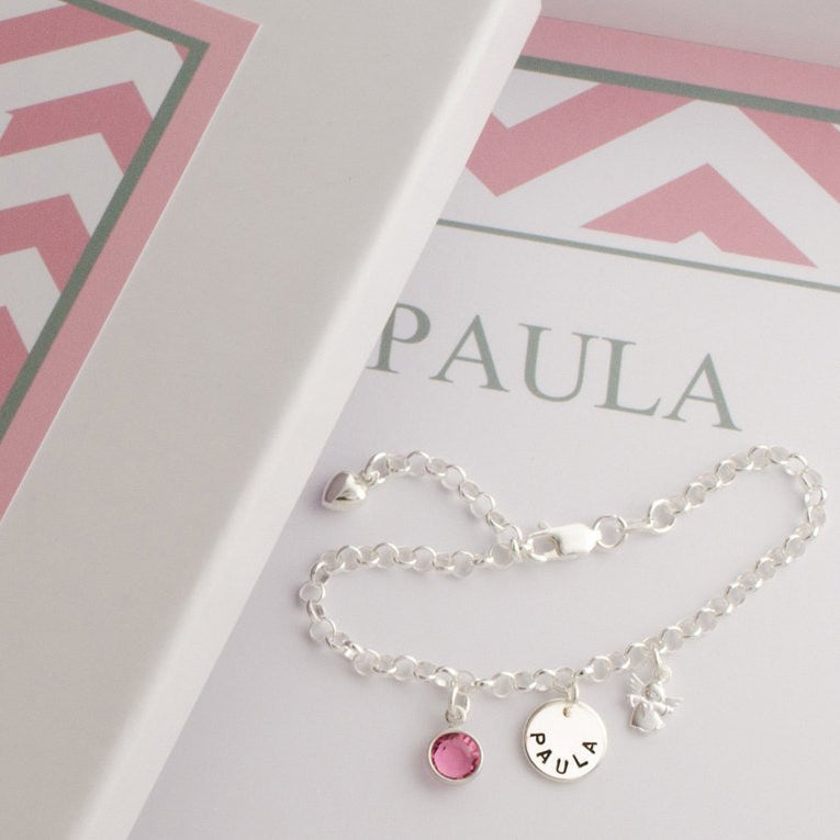 Detailansicht eines filigranen, verstellbaren Kinderarmbandes aus Sterlingsilber mit Engel, Namens-Anhänger mit Wunschgravur von Hand gestempelt und Geburtsstein. Das Armband liegt in einem Geschenkkarton, der mit Name und persönlichem Text versehen wurde.