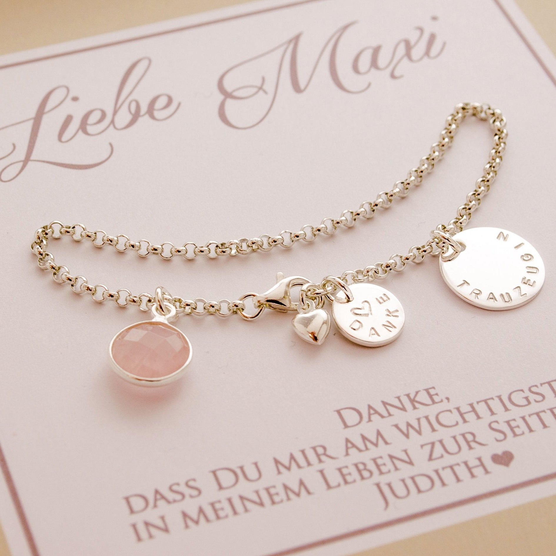 zierliches Gliederarmband mit Rosenquarz, kleinem Gravur-Taler mit 'Danke' und einem Herz mittig gestempelt und einem großen von Hand gestempelten Anhänger mit Gravur 'Trauzeugin'. Am Ende des Armbandes ist ein kleines bauchiges Herz befestigt. Das Armband wird in einer süßen personalisierten Geschenkbox in beige-weiß präsentiert.